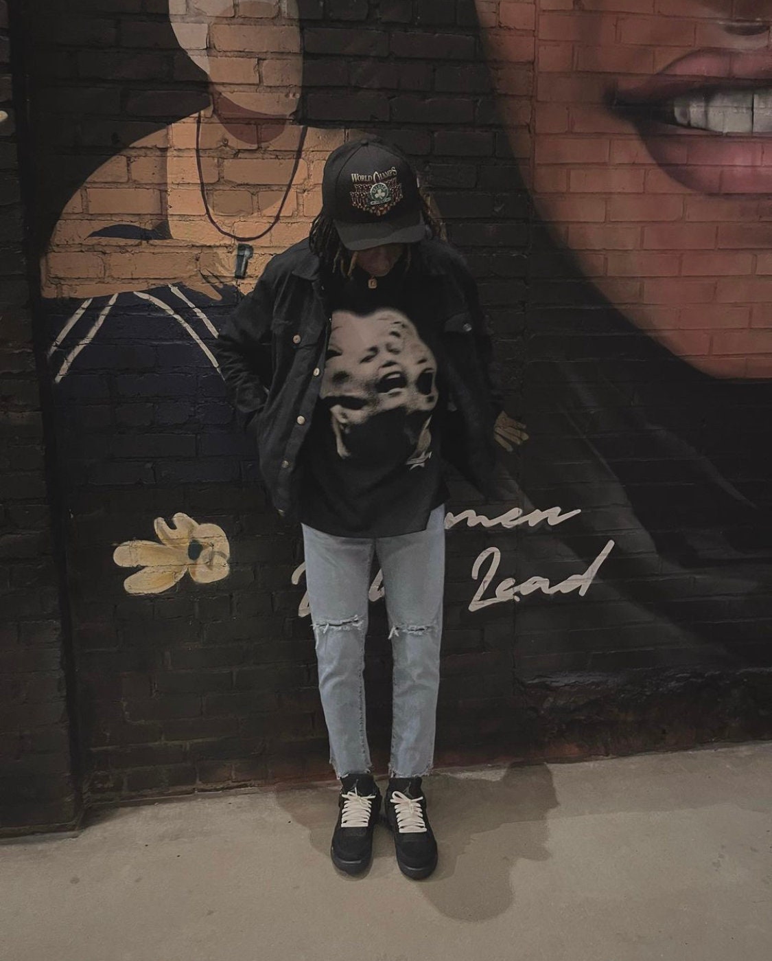 A person wearing a black jacket, a black t-shirt with a graphic print of three faces, blue jeans with distressed knees, and a black baseball cap with white text that reads 'World Order' and a graphic of a skull. The person is standing in front of a brick wall with a mural of a woman's face and the words 'Women Lead'.