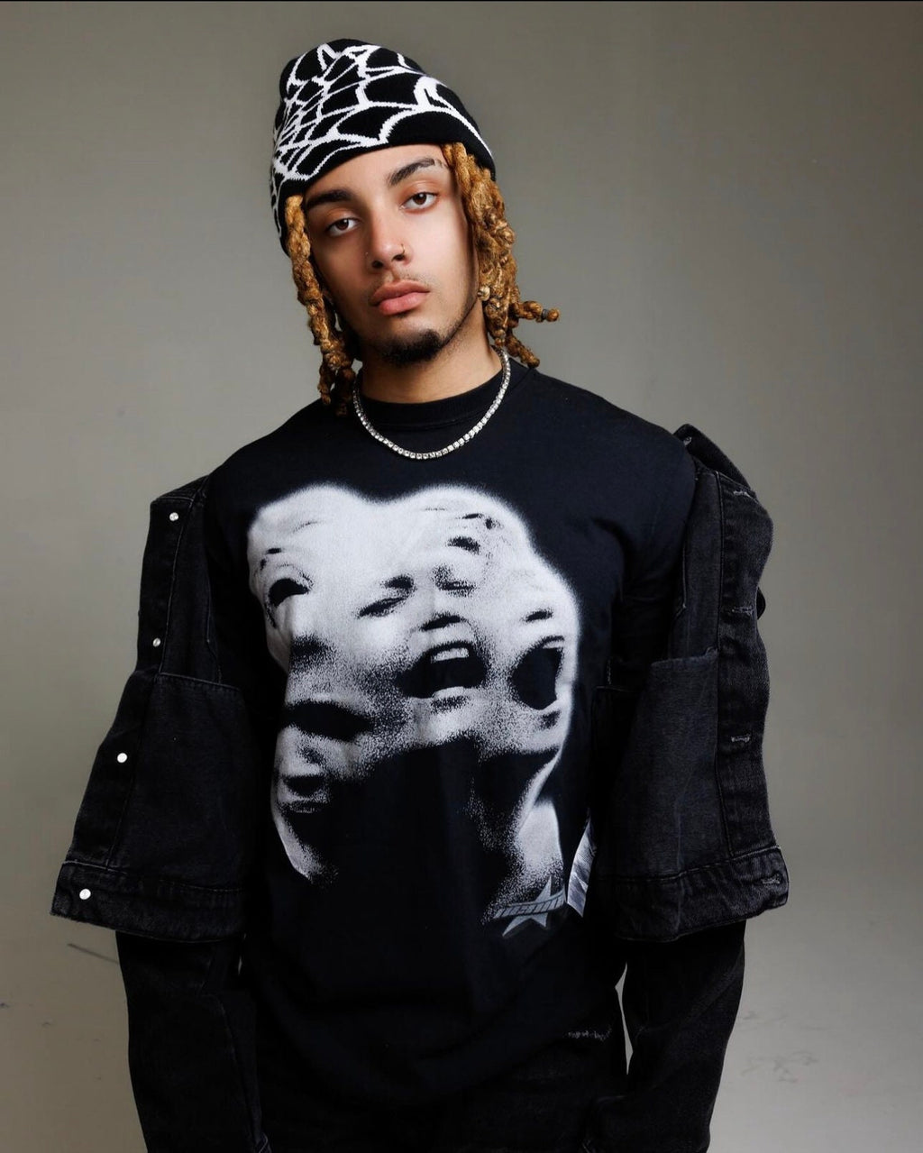 A person wearing a black long-sleeved shirt with a white graphic print of three faces, a black and white knit beanie, and a black denim jacket.