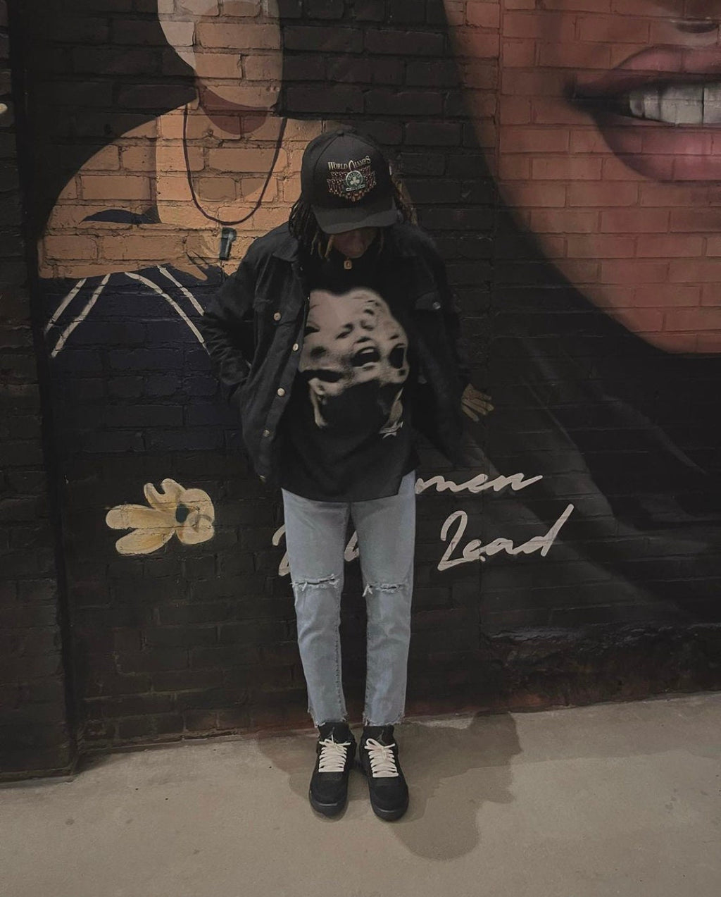 A person wearing a black jacket, a black t-shirt with a graphic print of three faces, blue jeans with distressed knees, and a black baseball cap with white text that reads 'World Order' and a graphic of a skull. The person is standing in front of a brick wall with a mural of a woman's face and the words 'Women Lead'.