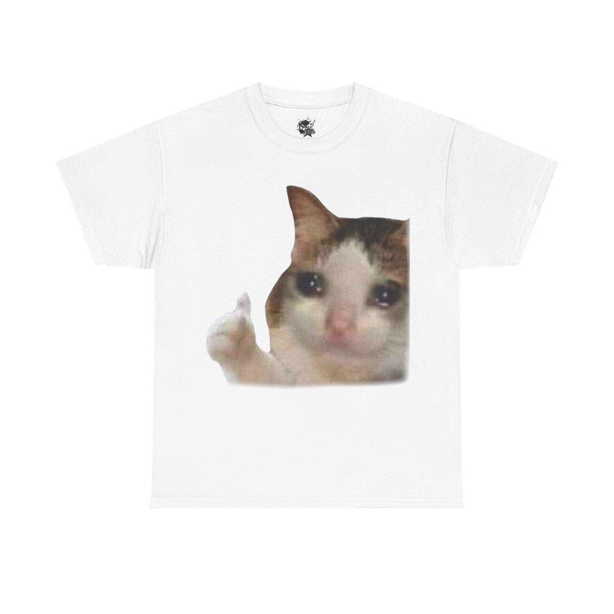 May include: A white t-shirt with a printed image of a cat with a sad expression and a single tear on its cheek. The cat is giving a thumbs up with its paw.
